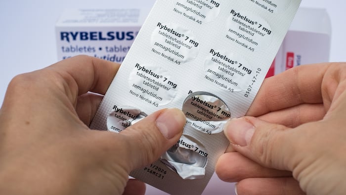 A box pills labelled Rybelsus in the background while a pair of hands pop a blister pack of Rybelsus to get a pill out