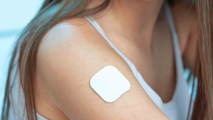 A white woman with brown long hair wears a white top and has a Nicotine patch on her right top arm