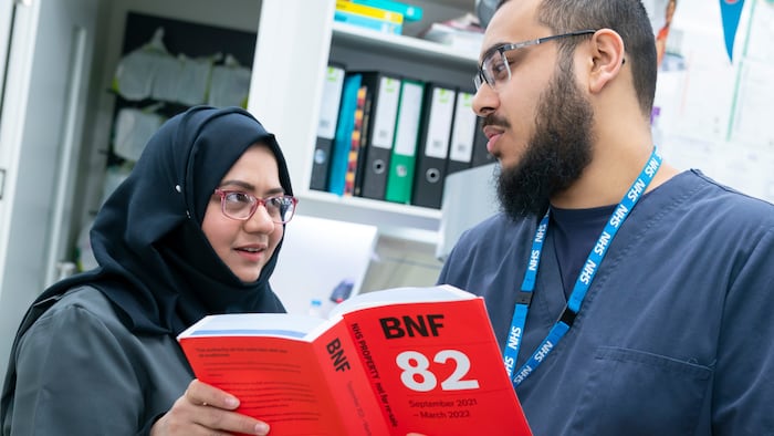 A Muslim woman wearing a Hijab looks at a brown man with a beard and both wear glasses. They are looking at each other while holding a red book titled 'BNF'.