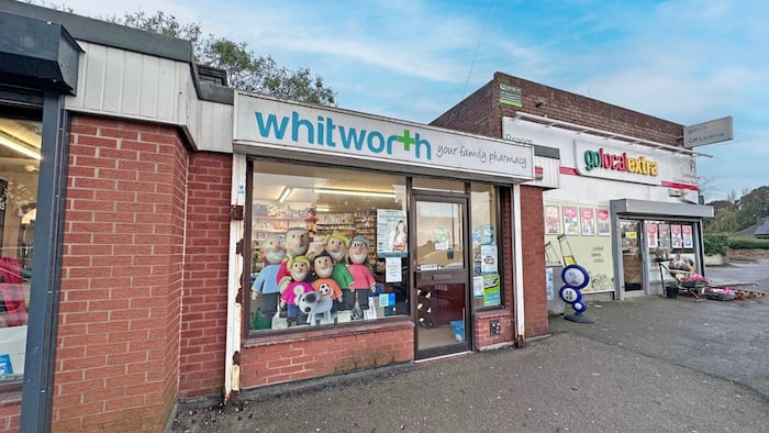 A pharmacy stands in a shopping parade next to a corner shop