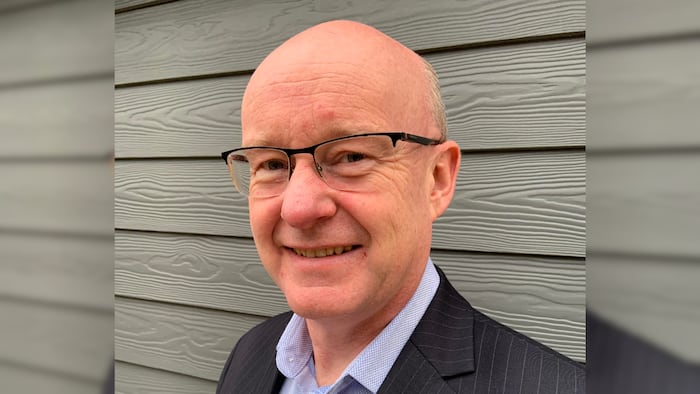 A bald white man wearing glasses and a pinstripe suit and light shirt smiling at the camera