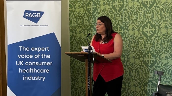 A white woman wearing a red top and glasses addresses a room at a stand giving a speech next to a poster that reads 'PAGB, the expert voice of the UK consumer healthcare industry'